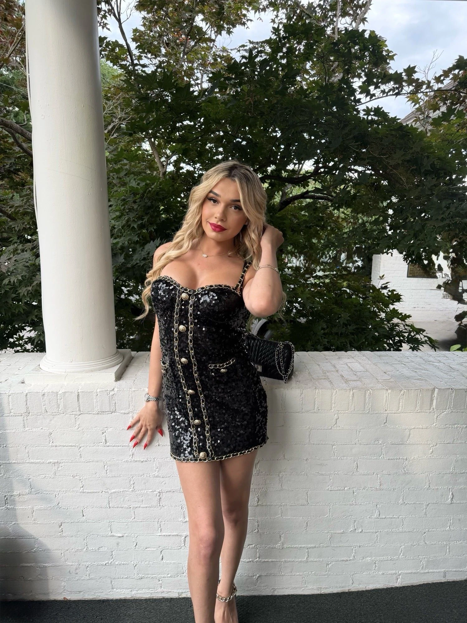 A black sequinned dress with gold chain detail, standing against white columns in evening light.